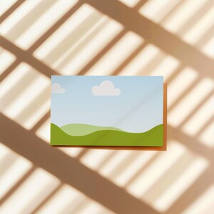 May include: A rectangular print featuring a landscape scene with rolling green hills under a blue sky with two white clouds. The print is set against a beige background with shadows.