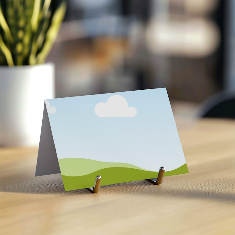 May include: A folded card with a landscape design, featuring a blue sky with white clouds and green rolling hills. The card is propped up by a metal stand on a wooden surface. A plant in a white pot is in the background.