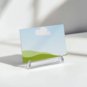 May include: A clear acrylic sign holder displaying a landscape image with a blue sky, white cloud, and green hills. The holder is angled and sits on a white surface, casting a shadow.