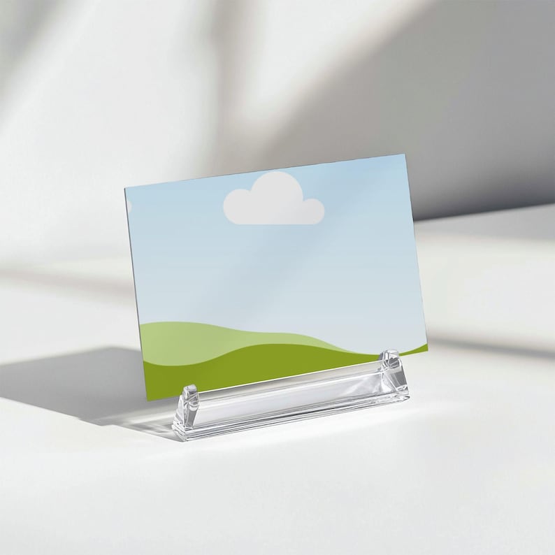 May include: A clear acrylic sign holder displaying a landscape image with a blue sky, white cloud, and green hills. The holder is angled and sits on a white surface, casting a shadow.