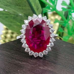 May include: A silver-toned ring featuring a large, oval-cut, deep red gemstone at the center. The central stone is surrounded by a halo of small, clear, round-cut white stones set in a scalloped metal frame, creating a classic cluster design.