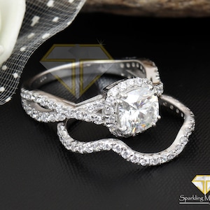 Peut inclure: Une bague de fiançailles argentée avec une pierre centrale taille coussin entourée d'un halo de petites pierres rondes. L'anneau présente un design torsadé incrusté de petites pierres, accompagné d'une alliance incurvée assortie.