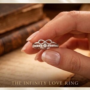 Puede incluir: Un anillo de plata con un diseño de símbolo de infinito, engastado con pequeñas piedras transparentes. El anillo se sostiene entre los dedos, con el texto "THE INFINITY LOVE RING" en la parte inferior de la imagen. El anillo es probablemente de plata de ley.