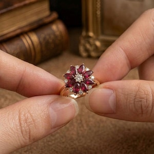 May include: A gold ring with a floral design. The flower is made up of deep red marquise-cut gemstones around a central diamond. Small white diamonds are set between the red gemstones. The ring is held between two fingers.