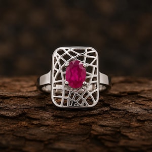 Op de afbeelding: Een zilverkleurige ring met een rechthoekig voorvlak voorzien van een ingewikkeld, opengewerkt web- of rasterpatroon. Een enkele, ovaal geslepen, diep roze-rode edelsteen is in het midden van het geometrische metalen frame gezet.