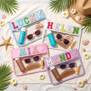 May include: Four clear vinyl pouches with colourful fabric letter patches spelling LUCKY, HELEN, MIA, and MANDY. Each pouch contains a bottle of Coola sunscreen, sunglasses, a wooden comb, and a lip balm, arranged on a white towel with seashells.