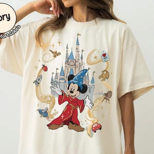 May include: Cream-coloured t-shirt with a graphic of Mickey Mouse dressed as a sorcerer, set against a castle backdrop and animated details. The shirt has the text "Ivory Comfort Colors" in a circle.