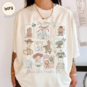 May include: Ivory-colored t-shirt featuring a colorful Toy Story graphic. The design includes various characters and elements from the movie, such as Woody, Buzz Lightyear, and a castle. The text "Toy Story" and "You've Got a Friend in Me" are also visible.