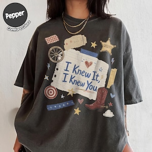 May include: A charcoal gray t-shirt featuring a central graphic of a lined notebook paper with the text "I Knew It, I Knew You" in blue. The design includes a red cowboy boot, a sheriff star, a wagon wheel, a target, and various small stars.