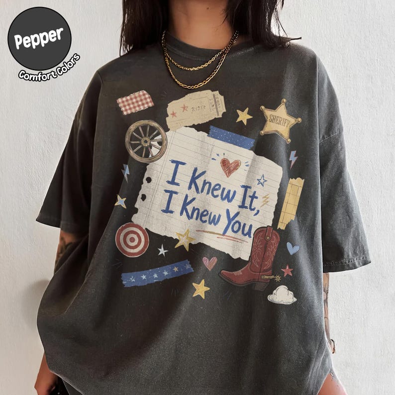 May include: A charcoal gray t-shirt featuring a central graphic of a lined notebook paper with the text "I Knew It, I Knew You" in blue. The design includes a red cowboy boot, a sheriff star, a wagon wheel, a target, and various small stars.