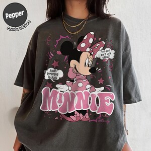 May include: Dark grey t-shirt featuring Minnie Mouse in a pink polka-dot dress and bow. The shirt has the word "MINNIE" in pink, along with the phrases "Always Remember To Laugh" and "Too-Hoo! Ain't Life Grand?" The shirt is from Disney.
