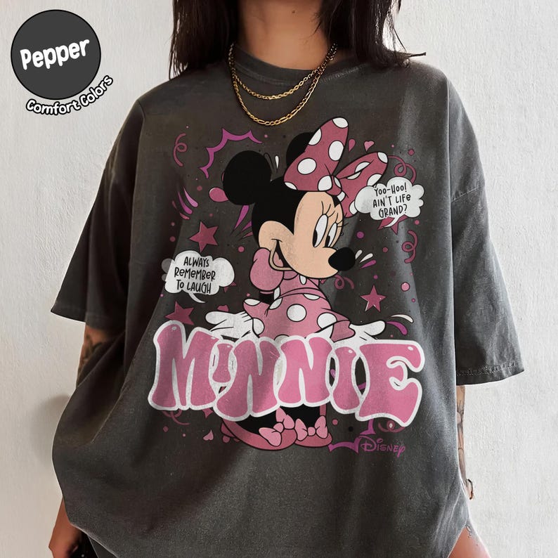 May include: Dark grey t-shirt featuring Minnie Mouse in a pink polka-dot dress and bow. The shirt has the word "MINNIE" in pink, along with the phrases "Always Remember To Laugh" and "Too-Hoo! Ain't Life Grand?" The shirt is from Disney.