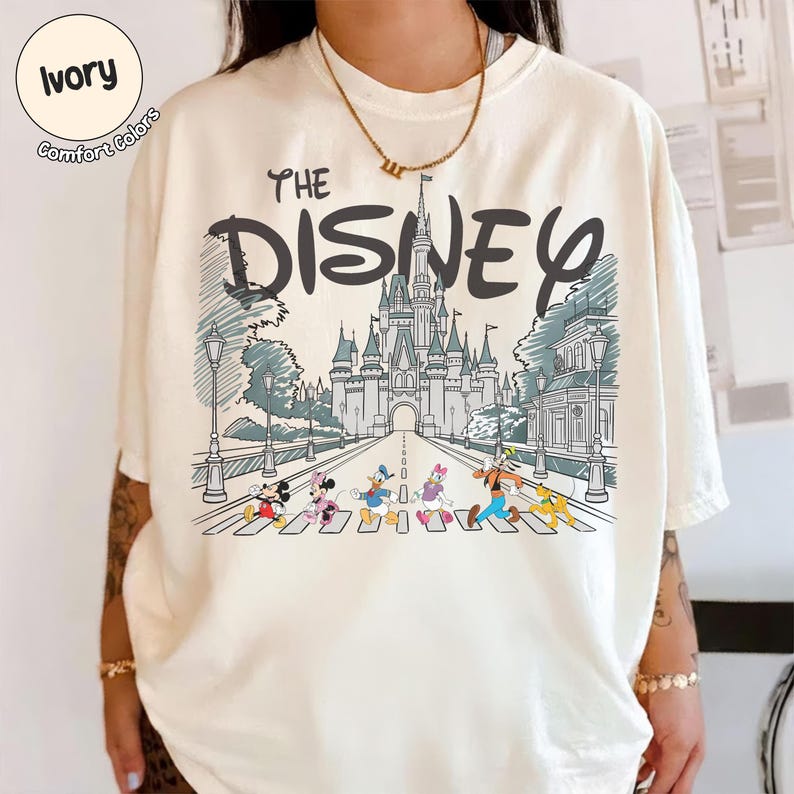 May include: An ivory t-shirt featuring a graphic print of a castle with the text "THE DISNEY" above it. Below the castle, a line of cartoon characters including Mickey Mouse, Minnie Mouse, Donald Duck, Daisy Duck, Goofy, and Pluto walk together.