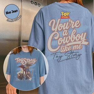 May include: A blue jean colored t-shirt featuring the Toy Story logo and the text "You're a Cowboy like me" on the back. The front graphic displays two figures with guitars and the text "Toy Story" in a stylized font with a blue brushstroke background.