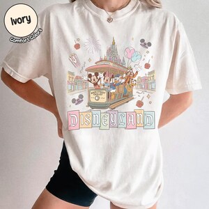 K&ouml;nnte beinhalten: Elfenbeinfarbenes T-Shirt mit einem Disneyland-Motiv. Die Grafik zeigt Micky Maus, Minnie Maus, Donald Duck, Goofy und Pluto in einer Main Street Trolley. Das Wort "Disneyland" wird in Pastellfarben unter der Stra&szlig;enbahn angezeigt.