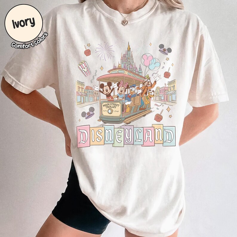 K&ouml;nnte beinhalten: Elfenbeinfarbenes T-Shirt mit einem Disneyland-Motiv. Die Grafik zeigt Micky Maus, Minnie Maus, Donald Duck, Goofy und Pluto in einer Main Street Trolley. Das Wort "Disneyland" wird in Pastellfarben unter der Stra&szlig;enbahn angezeigt.