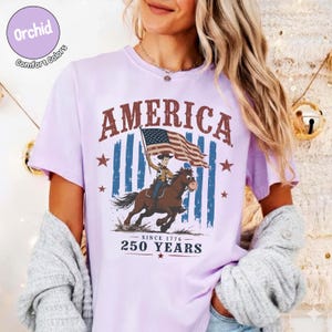May include: A light purple t-shirt featuring a graphic of a cowboy riding a horse while holding an American flag. The text reads AMERICA at the top and SINCE 1776 250 YEARS at the bottom. The design includes blue vertical stripes and red stars.
