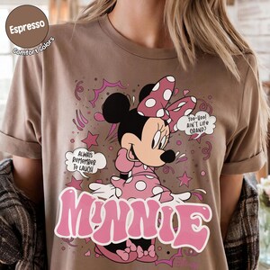 May include: Espresso-colored t-shirt featuring Minnie Mouse in a pink polka-dot dress and bow. The design includes the word "MINNIE" in pink, along with the phrases "Always Remember To Laugh" and "Yoo-Hoo! Ain't Life Grand?"