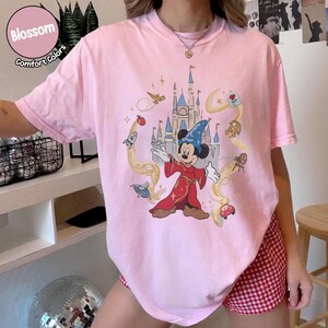 May include: A pale pink t-shirt with a graphic of Mickey Mouse as a sorcerer, set against a castle scene with animated details. The shirt includes the words "Blossom" and "Comfort Colors" in a circular design.