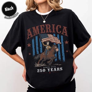 May include: A black t-shirt featuring a graphic of a cowboy riding a horse and holding an American flag. The text reads AMERICA at the top and SINCE 1776 250 YEARS at the bottom. A circular badge in the top left corner says Black Comfort Colors.