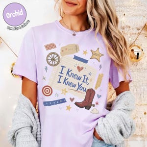 May include: A light purple short-sleeve t-shirt featuring a central graphic of a lined notepad page with the text I Knew It, I Knew You. The design includes a brown cowboy boot, a wagon wheel, a sheriff star, and various small decorative elements.