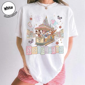 K&ouml;nnte beinhalten: Wei&szlig;es T-Shirt mit einem Main Street Trolley-Design mit Micky Maus, Minnie Maus, Donald Duck und Goofy. Das Wort "Disneyland" wird unterhalb der Stra&szlig;enbahn in bunten Buchstaben angezeigt. Das Shirt hat auch die W&ouml;rter "White" und "Comfort Colors".