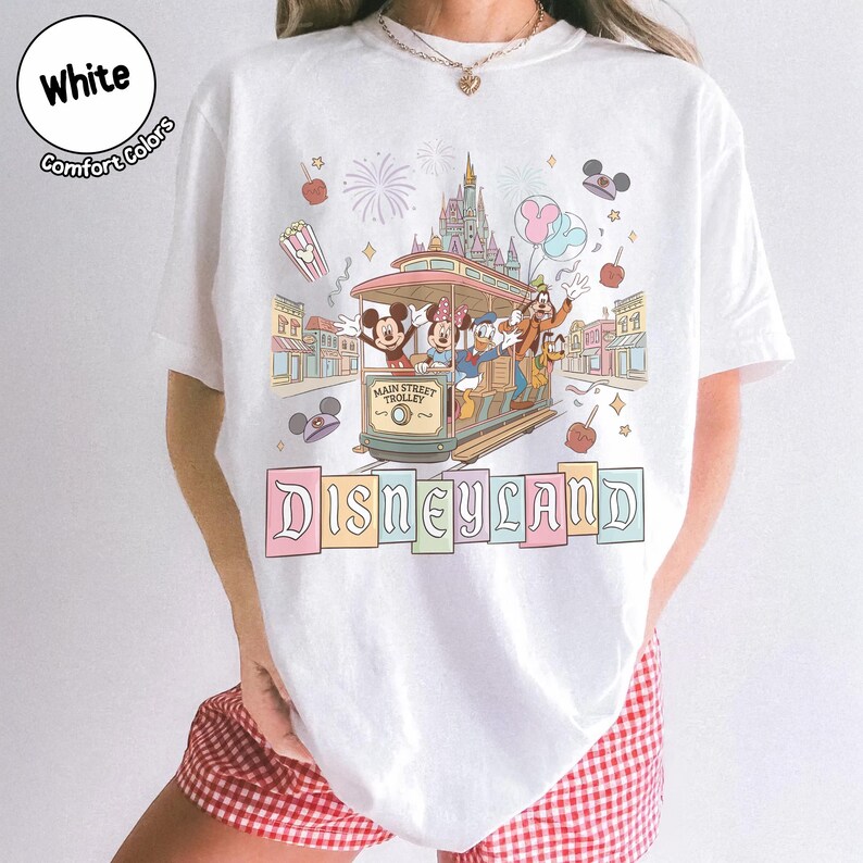 K&ouml;nnte beinhalten: Wei&szlig;es T-Shirt mit einem Main Street Trolley-Design mit Micky Maus, Minnie Maus, Donald Duck und Goofy. Das Wort "Disneyland" wird unterhalb der Stra&szlig;enbahn in bunten Buchstaben angezeigt. Das Shirt hat auch die W&ouml;rter "White" und "Comfort Colors".