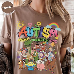 K&ouml;nnte beinhalten: Espressofarbenes T-Shirt mit einer bunten Grafik mit dem Wort "AUTISMUS" und den S&auml;tzen "akzeptieren, verstehen, lieben". Das Design umfasst Zeichentrickfiguren und einen Regenbogen.