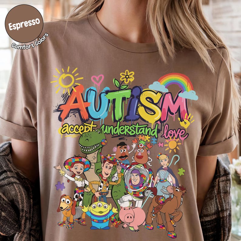 K&ouml;nnte beinhalten: Espressofarbenes T-Shirt mit einer bunten Grafik mit dem Wort "AUTISMUS" und den S&auml;tzen "akzeptieren, verstehen, lieben". Das Design umfasst Zeichentrickfiguren und einen Regenbogen.