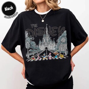 May include: A black t-shirt featuring a graphic print of a castle and Disney characters walking on a crosswalk. The text "THE DISNEY" is printed in a stylized font above the castle illustration. The shirt has a crew neckline and short sleeves.