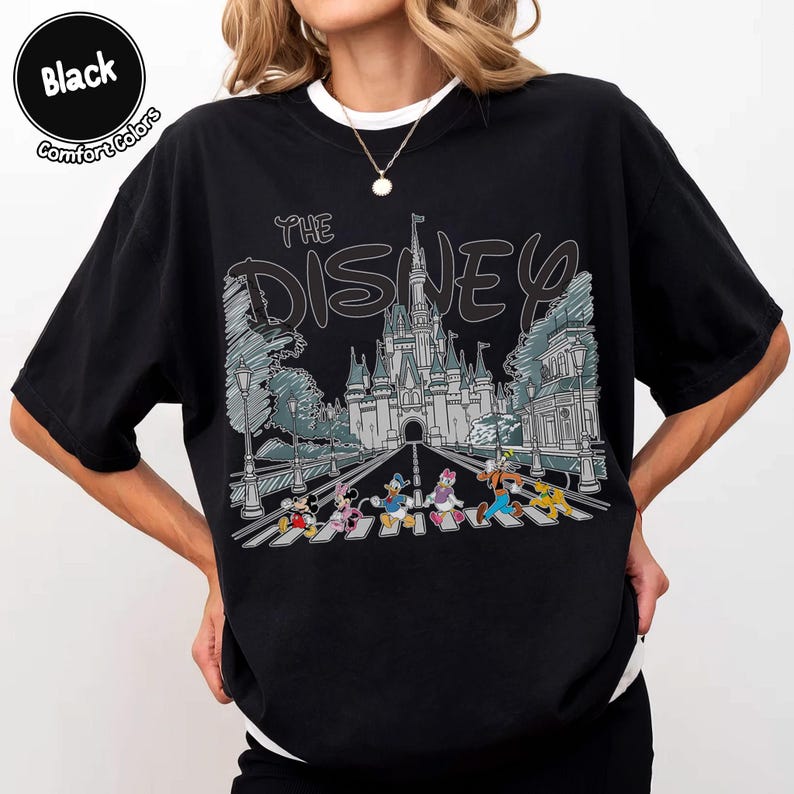 May include: A black t-shirt featuring a graphic print of a castle and Disney characters walking on a crosswalk. The text "THE DISNEY" is printed in a stylized font above the castle illustration. The shirt has a crew neckline and short sleeves.