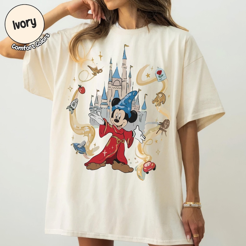 May include: Cream-coloured t-shirt with a graphic of Mickey Mouse dressed as a sorcerer, set against a castle backdrop and animated details. The shirt has the text "Ivory Comfort Colors" in a circle.