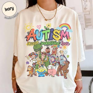 K&ouml;nnte beinhalten: Elfenbeinfarbenes T-Shirt mit einer bunten Grafik mit dem Wort "AUTISMUS" &uuml;ber den Worten "akzeptieren verstehen lieben". Die Grafik enth&auml;lt Zeichentrickfiguren und einen Regenbogen. Das Shirt ist aus Comfort Colors Material.