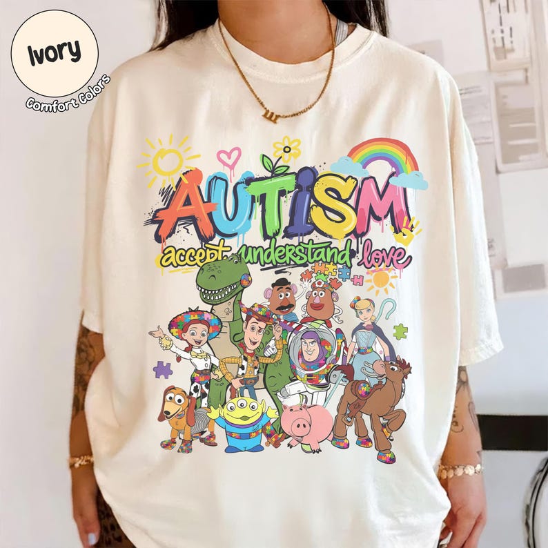 K&ouml;nnte beinhalten: Elfenbeinfarbenes T-Shirt mit einer bunten Grafik mit dem Wort "AUTISMUS" &uuml;ber den Worten "akzeptieren verstehen lieben". Die Grafik enth&auml;lt Zeichentrickfiguren und einen Regenbogen. Das Shirt ist aus Comfort Colors Material.
