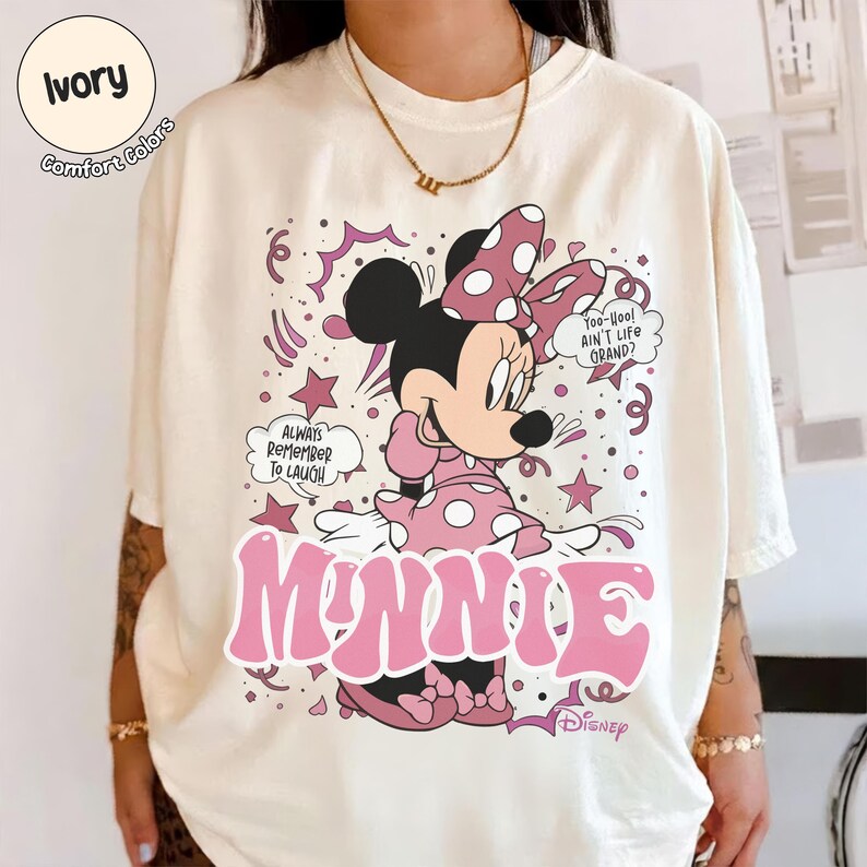 May include: Ivory-colored t-shirt featuring a graphic of Minnie Mouse in a pink polka-dot dress and bow. The design includes the word "MINNIE" in large pink letters, along with the phrases "Always Remember To Laugh" and "Yoo-hoo! Ain't Life Grand?"
