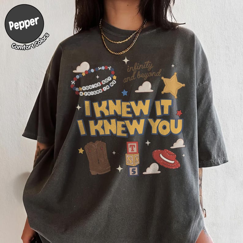 Puede incluir: Una camiseta de manga corta gris carb&oacute;n con un estampado gr&aacute;fico que incluye el texto I KNEW IT I KNEW YOU, infinity and beyond, y YOU'VE GOT A FRIEND IN ME. El dise&ntilde;o muestra una pulsera de cuentas, una estrella de sheriff, botas, bloques y un sombrero.