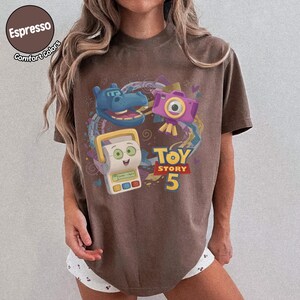 May include: A brown short-sleeve t-shirt featuring a graphic print of a blue hippopotamus, a purple camera, and a white toy device. The text "TOY STORY 5" is printed in yellow and red. The shirt has a crew neckline and a relaxed fit.