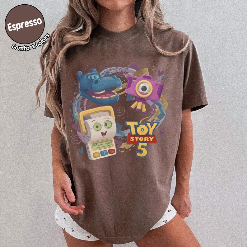May include: A brown short-sleeve t-shirt featuring a graphic print of a blue hippopotamus, a purple camera, and a white toy device. The text "TOY STORY 5" is printed in yellow and red. The shirt has a crew neckline and a relaxed fit.
