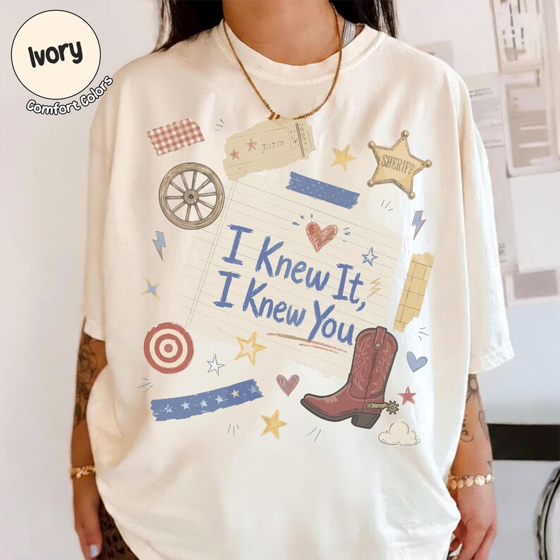 May include: An ivory t-shirt featuring a central graphic on lined paper with the text "I Knew It, I Knew You" in blue script. The design includes a brown cowboy boot, a wagon wheel, a sheriff star, and various small stars, hearts, and stripes.
