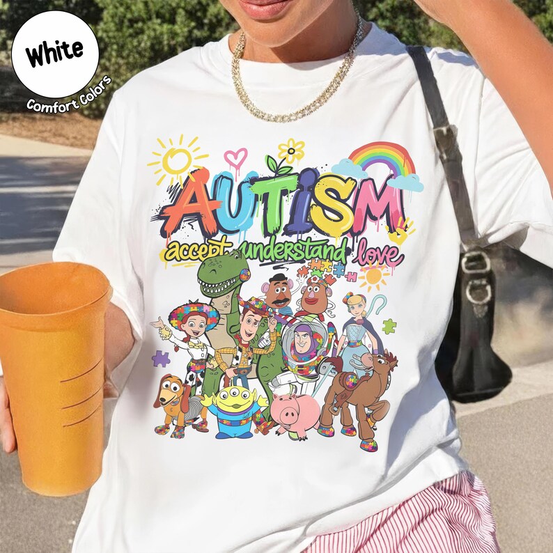 K&ouml;nnte beinhalten: Wei&szlig;es T-Shirt mit einer bunten Grafik mit dem Wort "AUTISMUS" und dem Satz "akzeptieren verstehen lieben". Das Design beinhaltet Zeichentrickfiguren und einen Regenbogen. Das Shirt hat die W&ouml;rter "Wei&szlig;" und "Comfort Colors" oben links.