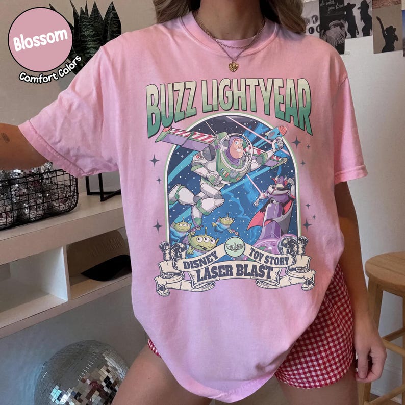 Puede incluir: Camiseta rosa claro con un dise&ntilde;o de Buzz Lightyear de Toy Story. El dise&ntilde;o incluye el texto "Buzz Lightyear" en verde y "Disney Laser Blast" en una pancarta. La camiseta tiene mangas cortas y cuello redondo.