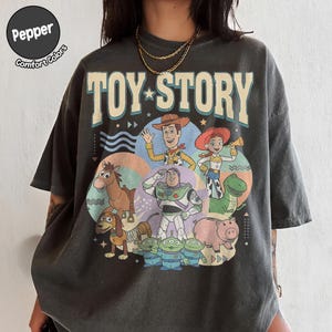 May include: Dark grey t-shirt with the words "TOY STORY" and a colourful graphic of characters from the animated film. The graphic includes Woody, Buzz Lightyear, Jessie, and other characters. The shirt is a comfort colours style.
