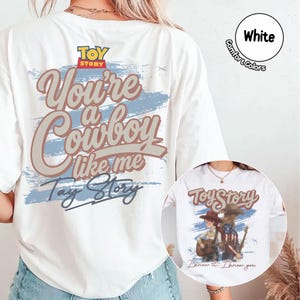 May include: A white t-shirt featuring a large graphic on the back with the text "Toy Story" and "You're a Cowboy like me Tay Story" in brown and blue. A circular inset shows the same shirt with a graphic of two figures and a guitar.