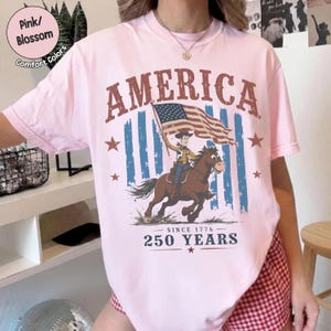 May include: A light pink t-shirt featuring a graphic of a cowboy riding a horse while holding an American flag. The text reads AMERICA at the top and SINCE 1776 250 YEARS at the bottom. The design includes blue vertical stripes behind the horse.