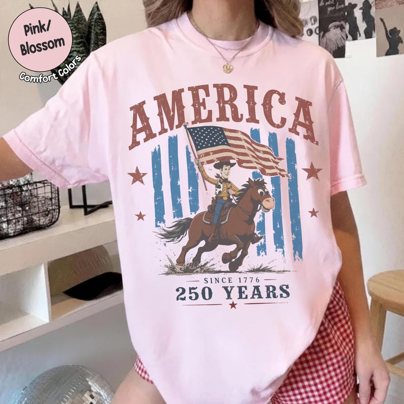 May include: A light pink t-shirt featuring a graphic of a cowboy riding a horse while holding an American flag. The text reads AMERICA at the top and SINCE 1776 250 YEARS at the bottom. The design includes blue vertical stripes behind the horse.