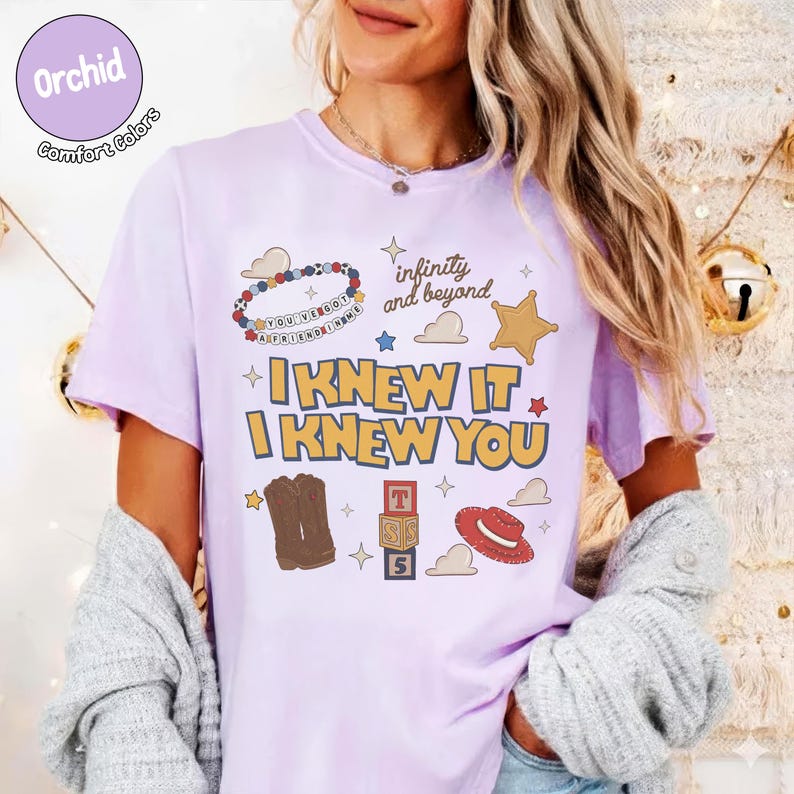 Puede incluir: Una camiseta de color morado claro con un estampado gr&aacute;fico que incluye el texto "I KNEW IT I KNEW YOU", "infinity and beyond" y "YOU'VE GOT A FRIEND IN ME". El dise&ntilde;o presenta ilustraciones de una pulsera de cuentas, una estrella, botas vaqueras, bloques y un sombrero.