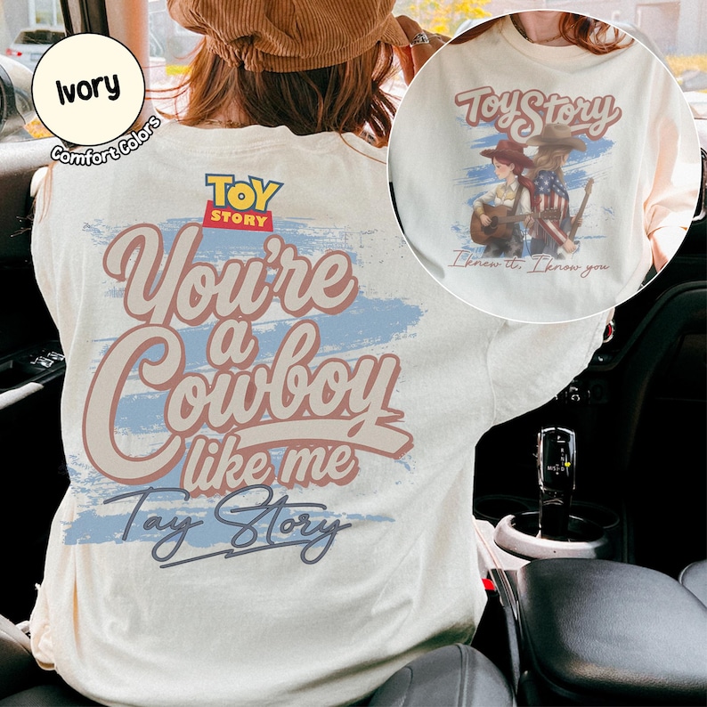 May include: An ivory t-shirt featuring a graphic print with the Toy Story logo and the text "You're a Cowboy like me" and "Tay Story" over a blue brushstroke. The back design includes a circular inset showing two figures with guitars.