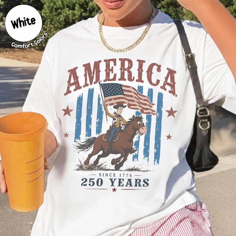 May include: A white t-shirt featuring a graphic of a cartoon cowboy riding a brown horse while holding an American flag. The text reads AMERICA, SINCE 1776, and 250 YEARS. The design includes blue vertical stripes and red stars on a white background.