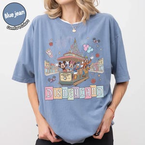 K&ouml;nnte beinhalten: Blaues Jeans-Comfort-Farben-T-Shirt mit einem Disneyland Main Street Trolley-Design mit Micky Maus, Minnie Maus, Donald Duck und Goofy. Das Shirt hat einen Rundhalsausschnitt und kurze &Auml;rmel. Das Wort "Disneyland" ist in Pastellfarben gedruckt.