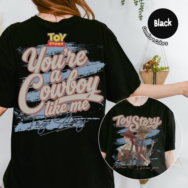 May include: A black t-shirt featuring a graphic print with the text "Toy Story" and "You're a Cowboy like me" in a vintage-style font. The design includes a blue brushstroke background and an illustration of two figures with guitars.
